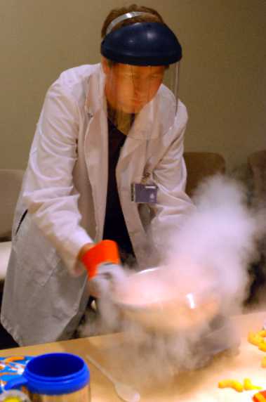 A person making liquid nitrogen ice cream dressed  in a lab coat with face shield. A bowl is emitting dry-ice-like smoke.